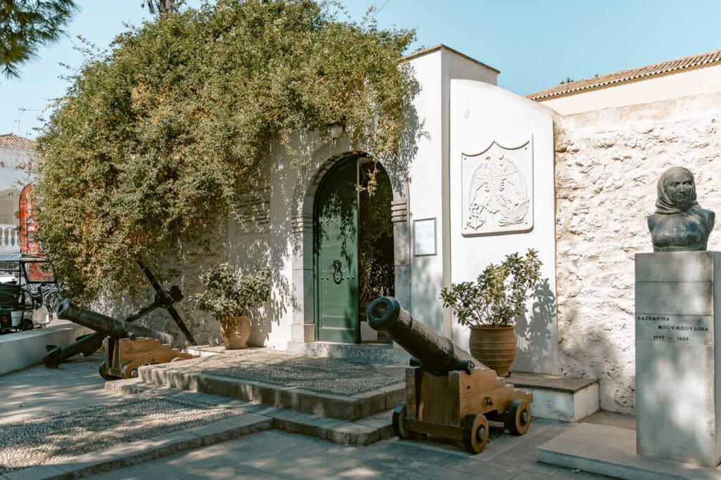 Entrance to the Bouboulina Museum in Spetses, Greece, with historic cannons, a green doorway, and a bronze bust outside.