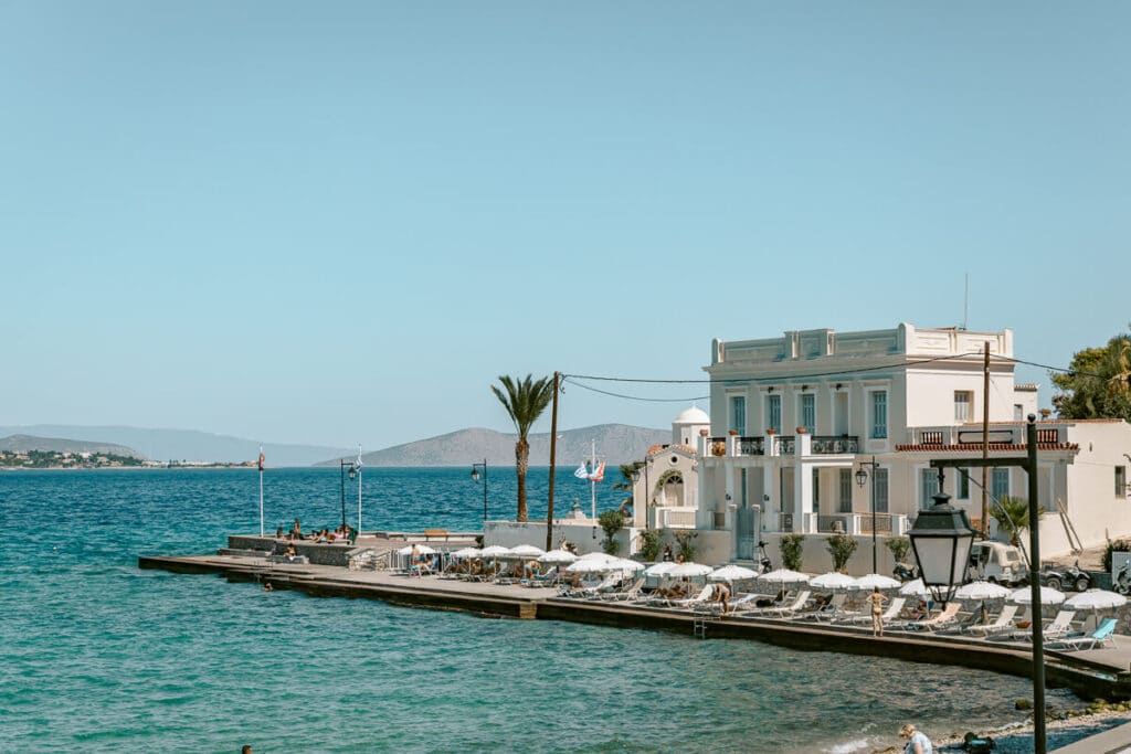 White waterfront building beside turquoise water in Spetses, with sun loungers and umbrellas lining the seaside terrace.