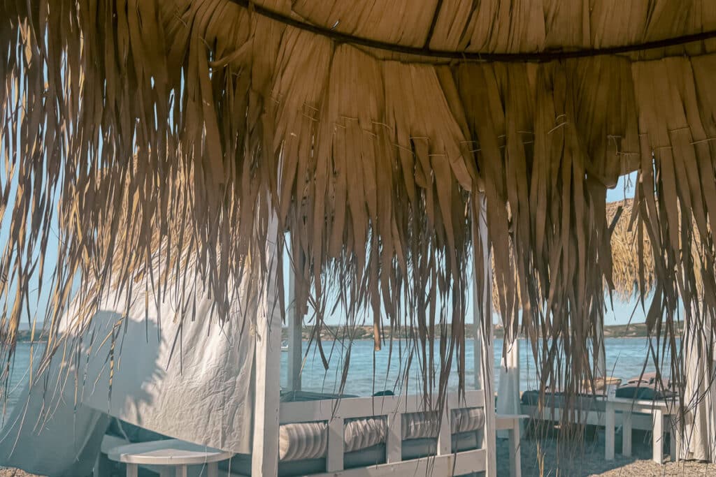 Shaded beach seating beneath a rustic straw umbrella overlooking the blue sea at Kaiki Beach in Spetses.