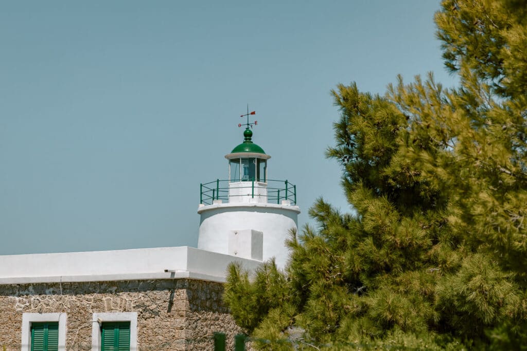 Spetses Lighthouse with its white tower and green roof rising above a stone building and surrounding pine trees.