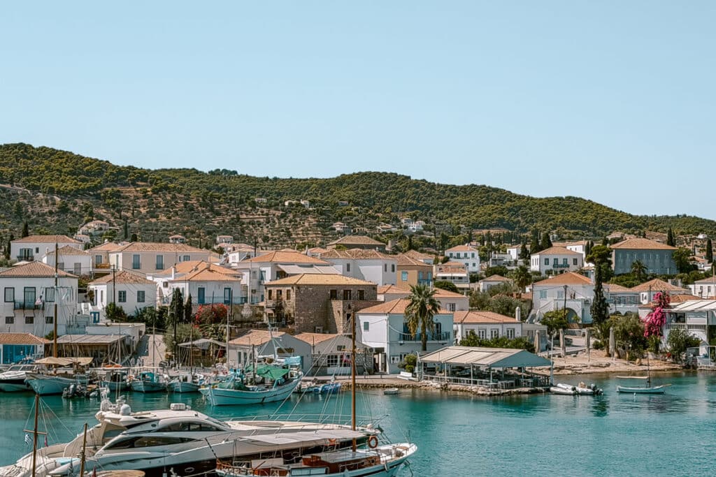 View of Spetses town from the harbor, with boats on turquoise water, white houses with terracotta roofs, and pine-covered hills in the background.