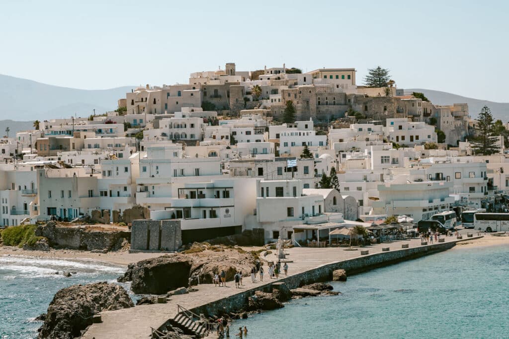 Whitewashed buildings and historic stone structures of Naxos Town, Greece, overlooking a waterfront walkway with people strolling beside the turquoise sea.