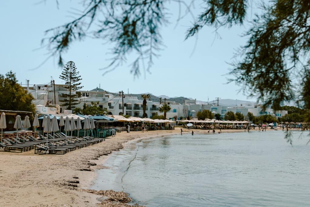 Agios Georgios Beach in Naxos, Greece, with shallow, calm water, sunbeds and umbrellas along the sandy shore, and white buildings in the background.