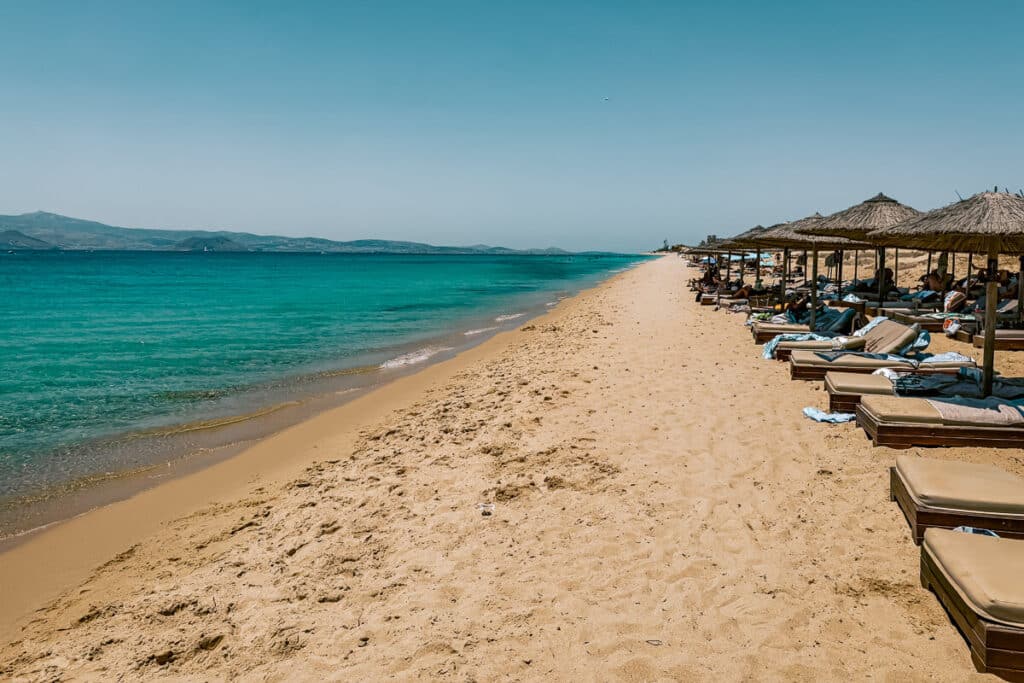 Maragkas Beach in Naxos, Greece, with golden sand, clear turquoise water, and rows of cushioned sun beds beneath thatched umbrellas.