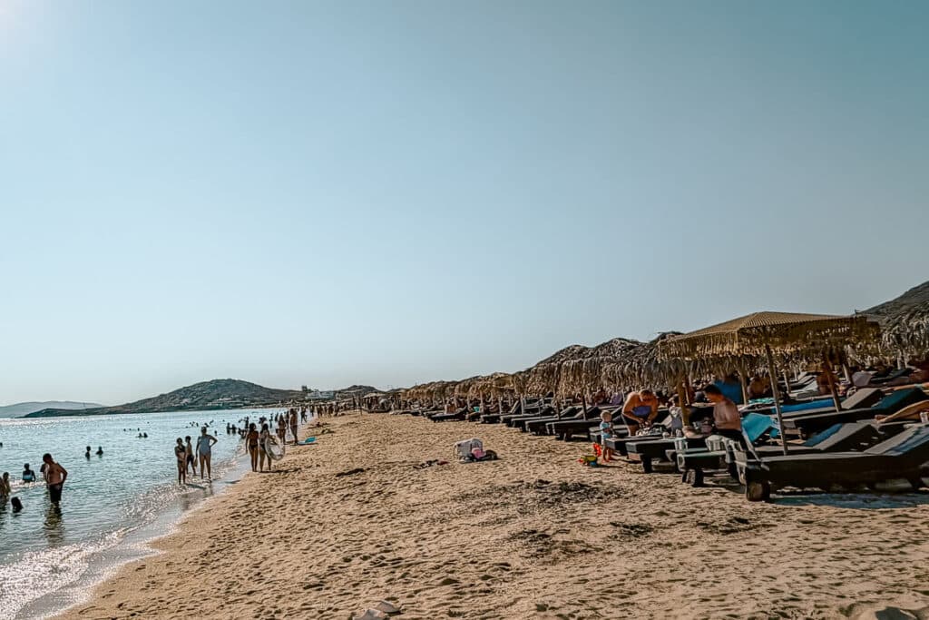 Agios Prokopios Beach in Naxos, Greece, crowded with swimmers along the sandy shoreline and rows of sunbeds beneath thatched umbrellas.