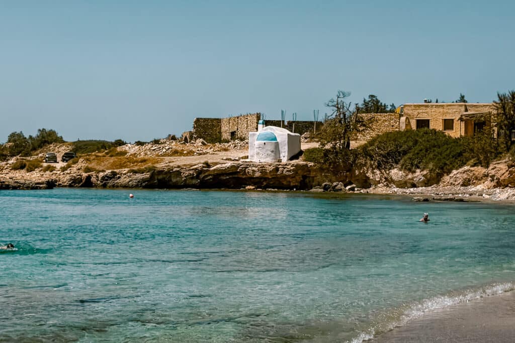 Alyko Beach in Naxos, Greece, with clear turquoise water, a swimmer, rocky shoreline, and traditional stone buildings overlooking the cove.