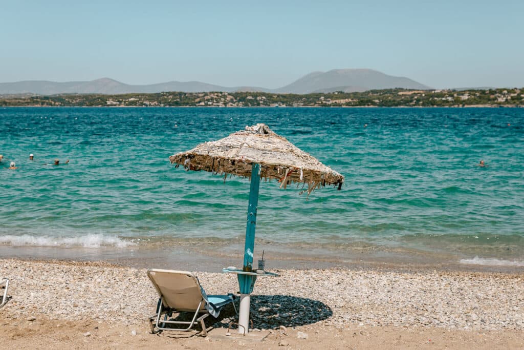 Straw beach umbrella and lounge chair on a pebbled beach overlooking turquoise water in Spetses, Greece.